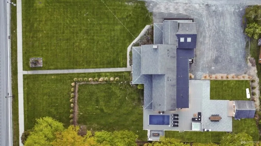 An aerial view shows a house beside lawns, paved areas, and surrounding trees.