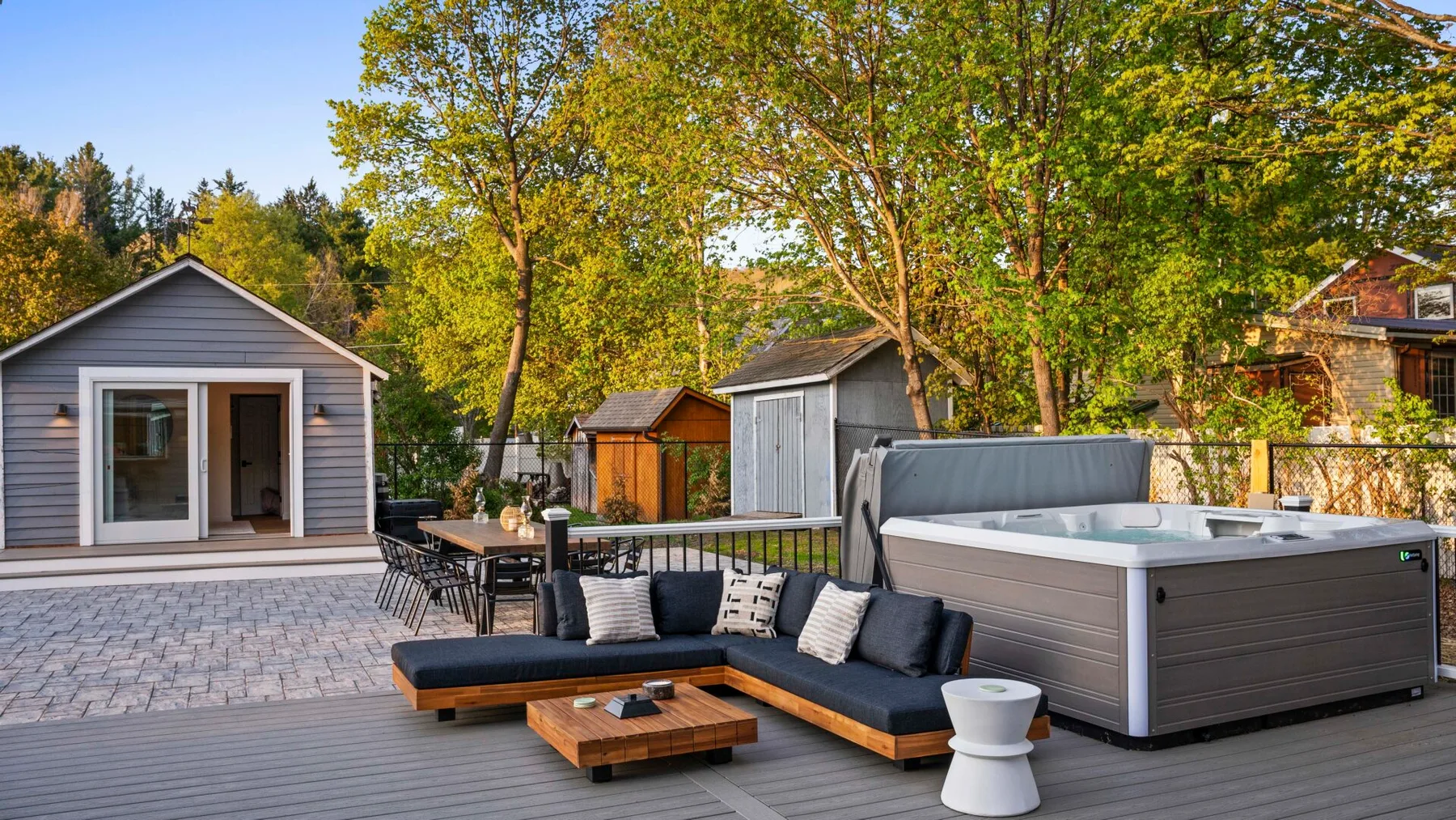 An outdoor patio has sectional seating, a hot tub, and small garden sheds.