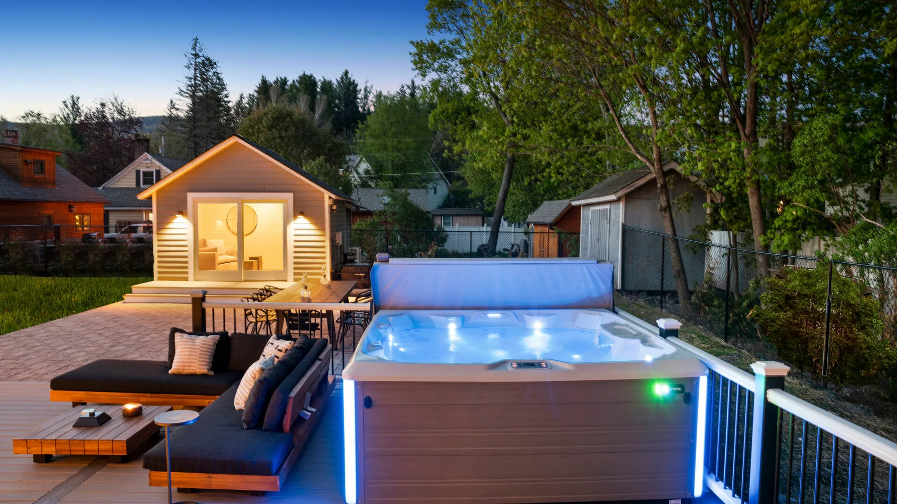 An outdoor hot tub glows beside lounge seating in a landscaped yard at dusk.