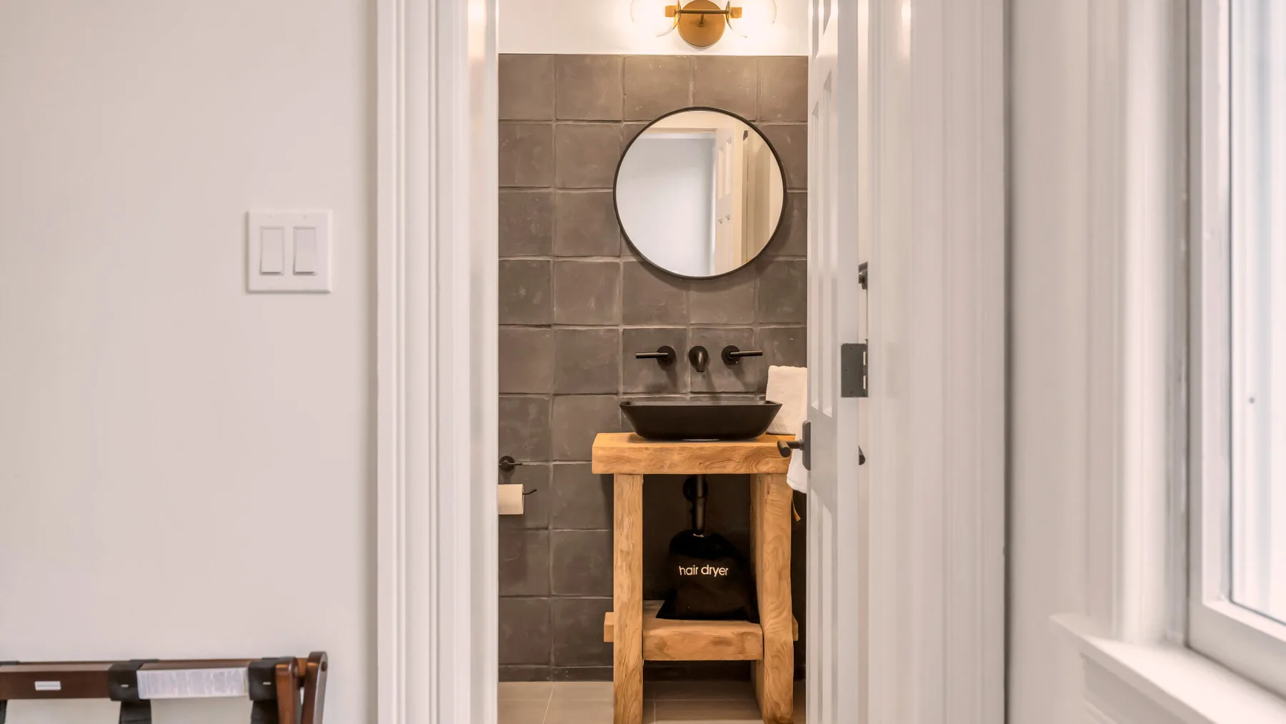 A bathroom vanity with a round mirror and dark tiled wall.