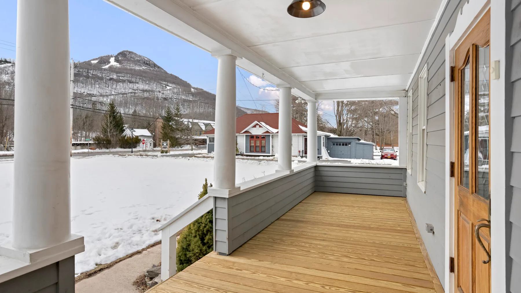 A covered porch with wooden flooring overlooks a snowy landscape and mountain.