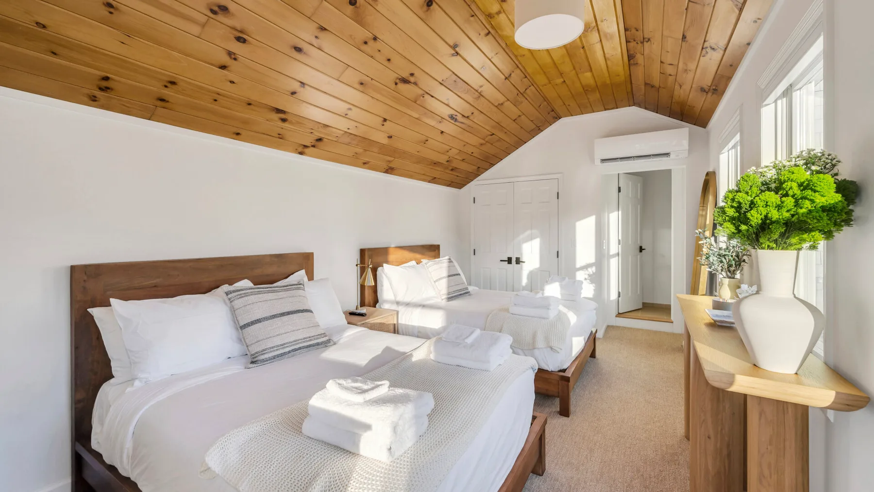 A bright bedroom with white bedding, a wooden ceiling, and large windows.