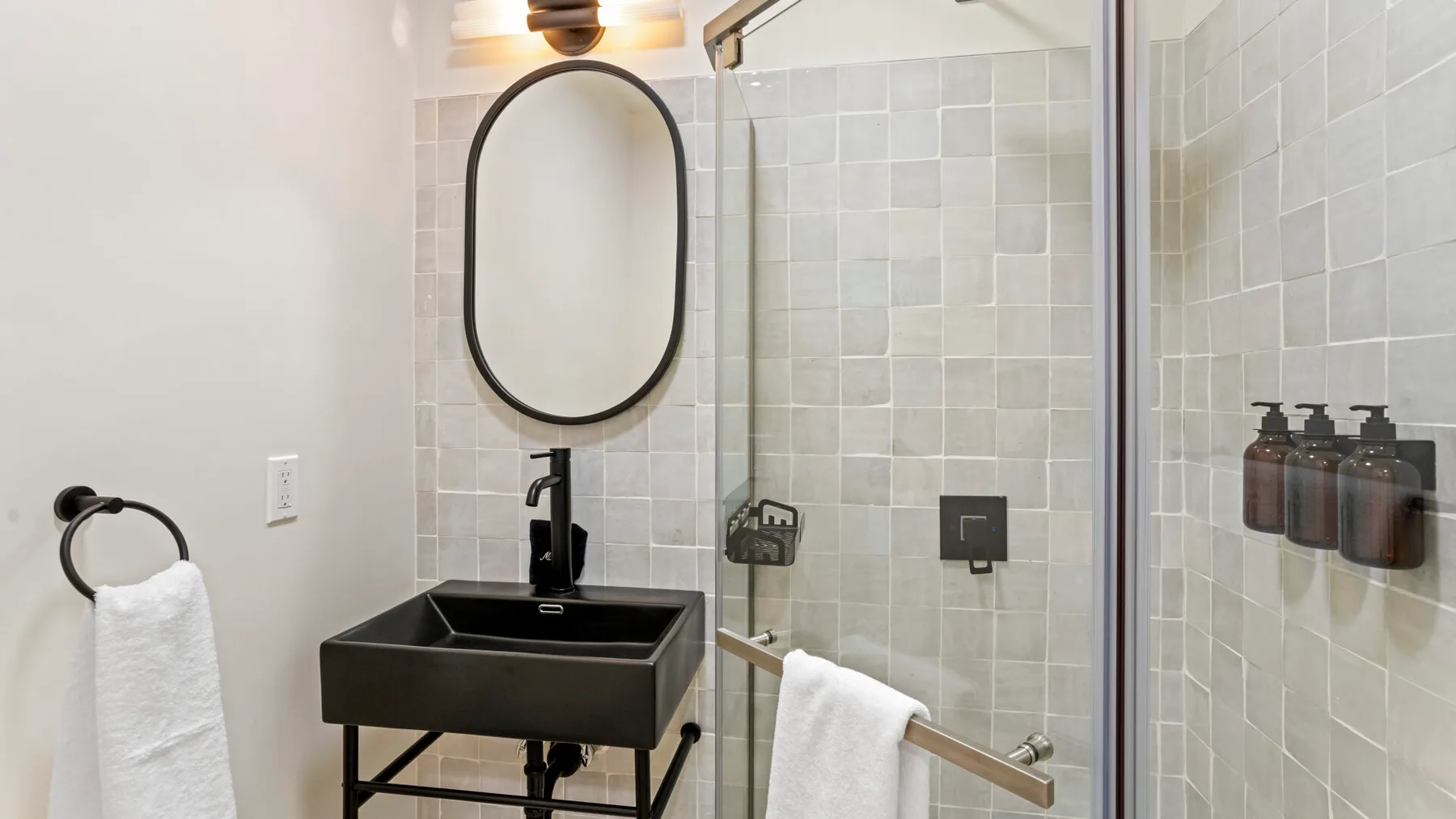 A tiled bathroom with a dark sink, oval mirror, and glass shower.