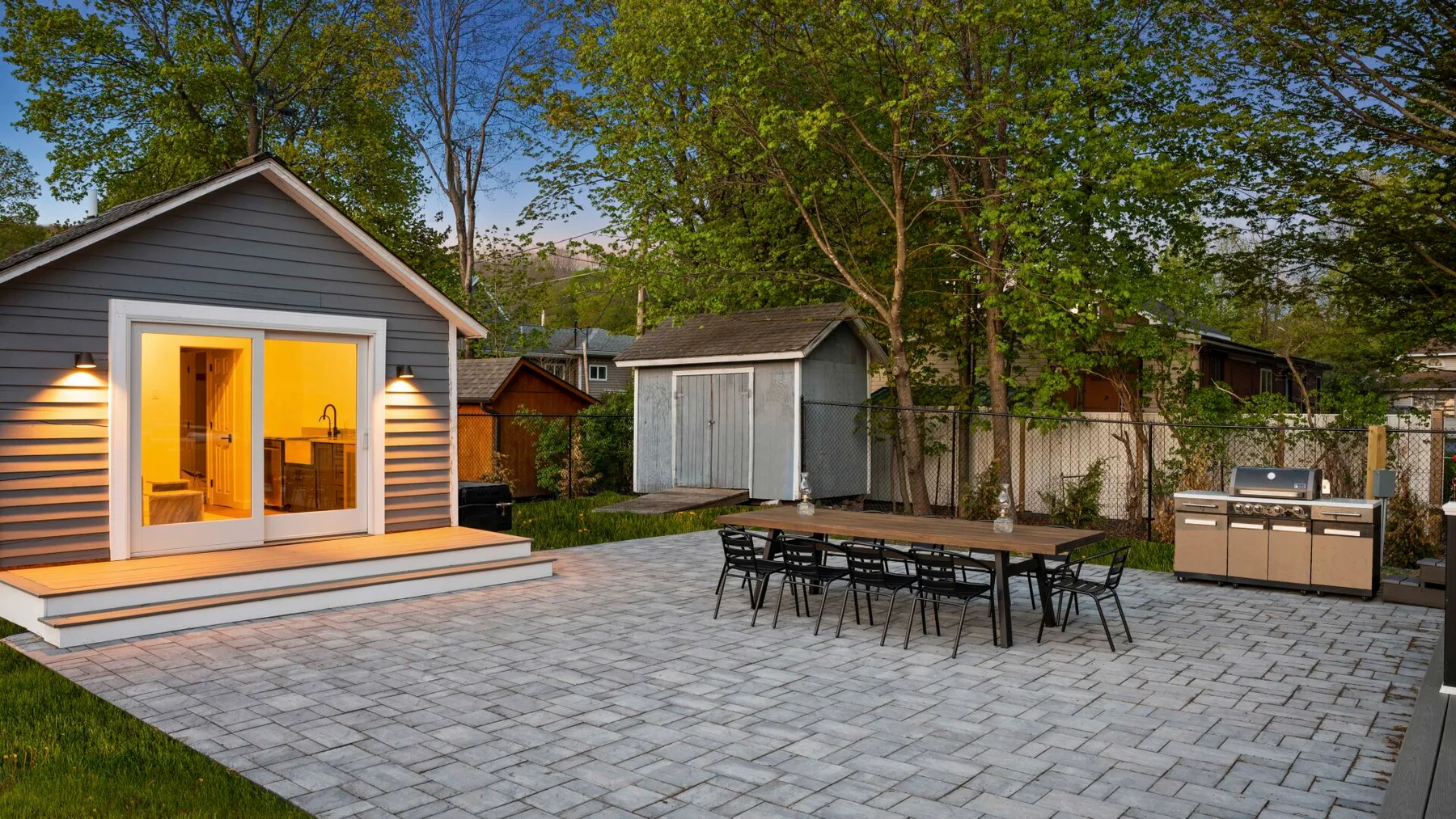 A paved outdoor patio has a dining table, seating, and a small shed.