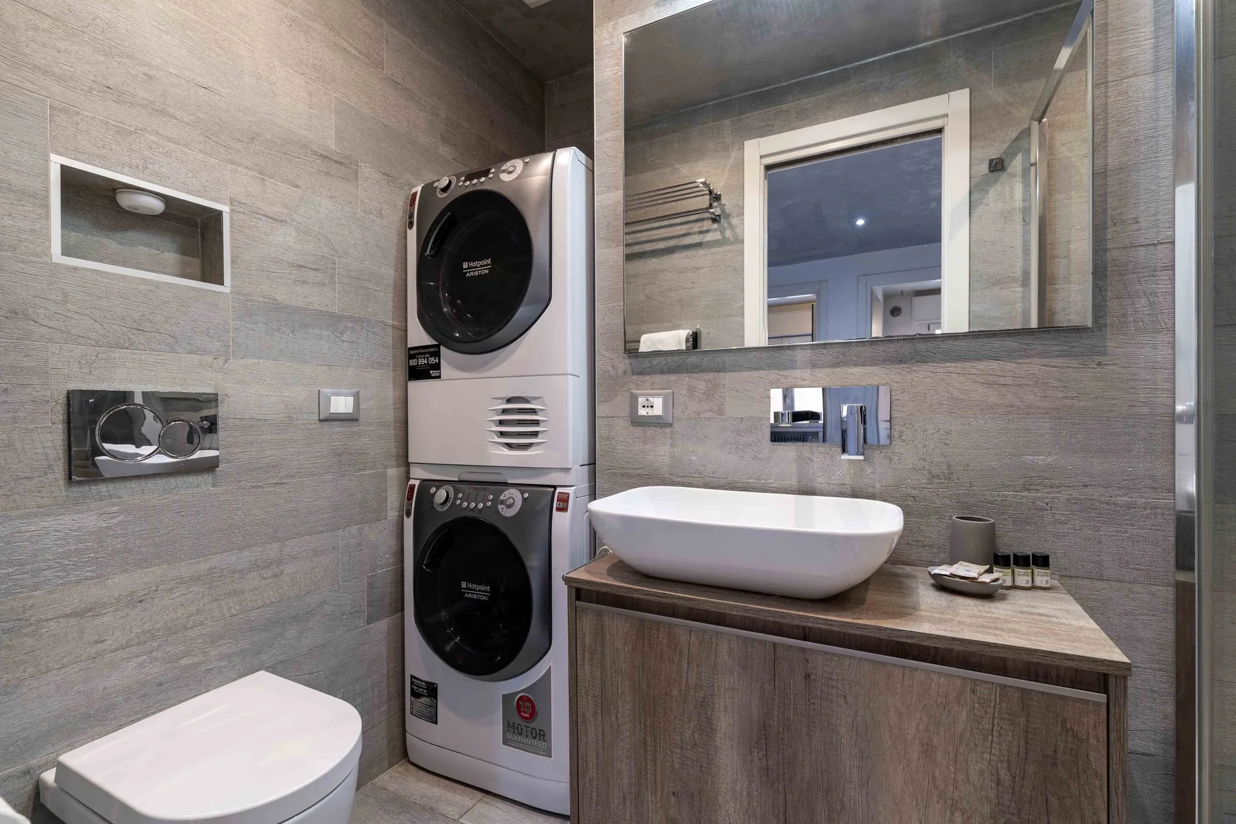 Baño revestido de azulejos con tocador, espejo y electrodomésticos de lavandería apilados.
