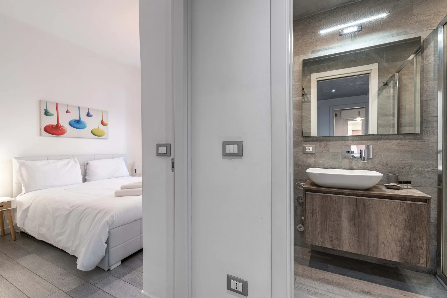 Un dormitorio con ropa de cama blanca aparece junto a un lavabo y un espejo.