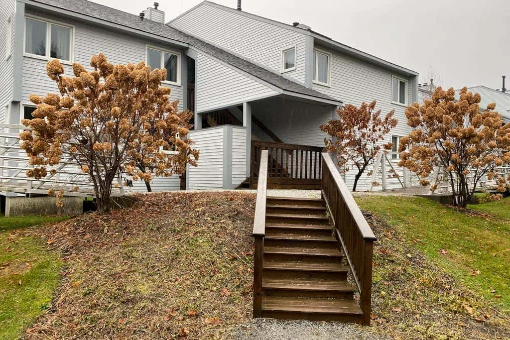 A covered entrance with wooden steps leads up to a light-colored house.