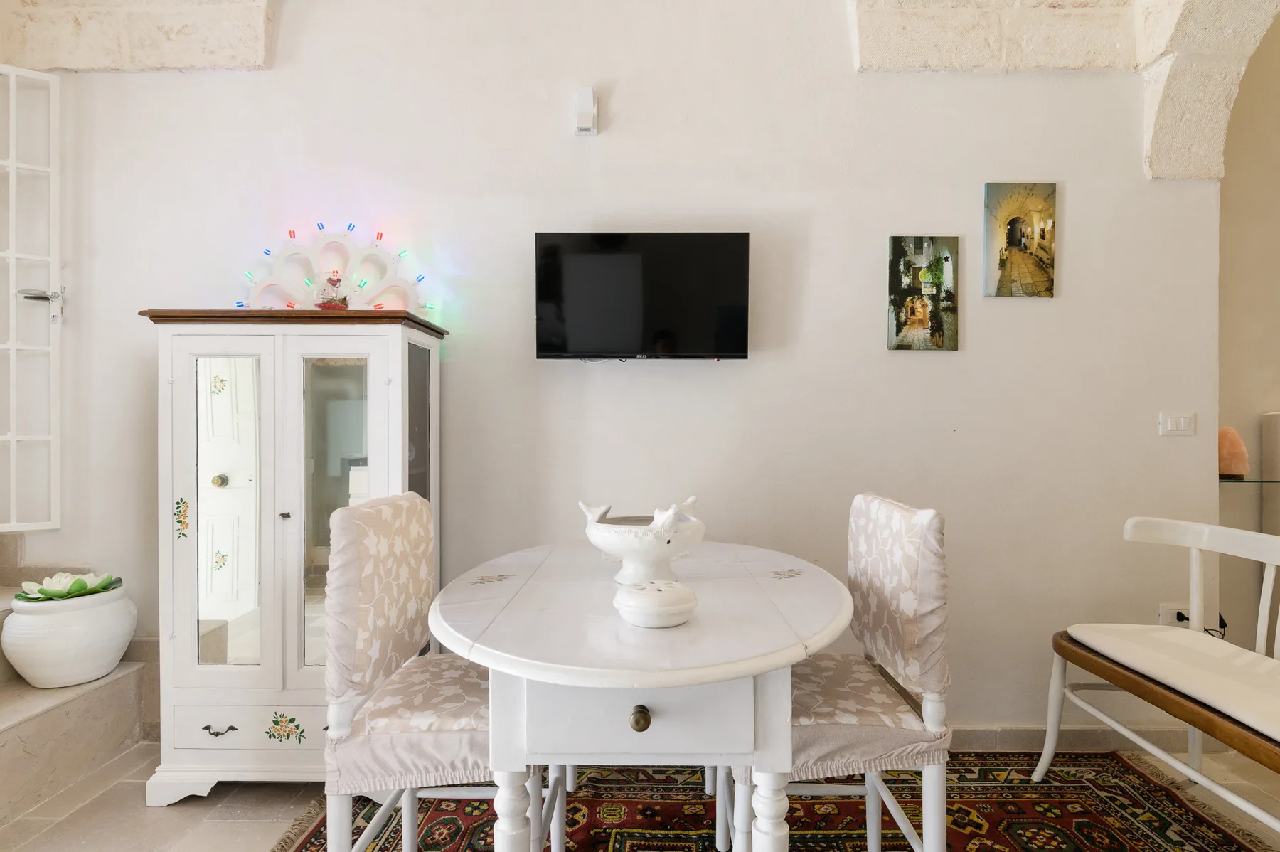 A bright dining area with a round table, upholstered seating, and a wall-mounted television.