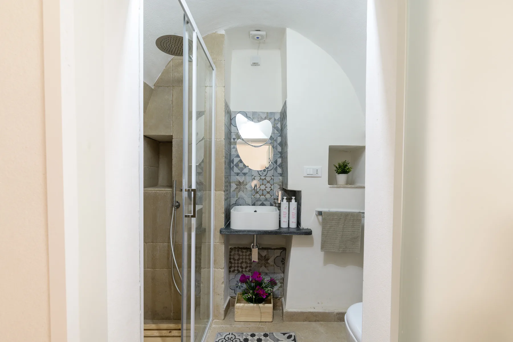 A bright bathroom with a glass shower and a sink beneath patterned tiles.