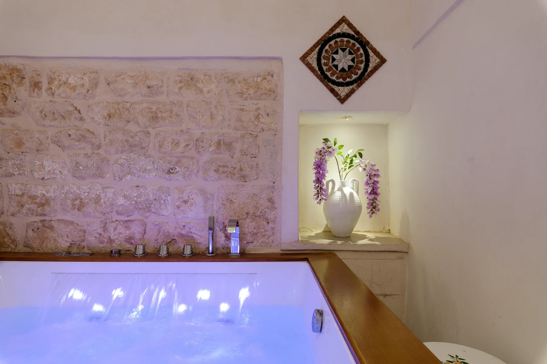 A lit indoor pool sits beneath a stone wall, with a flower arrangement in a niche.