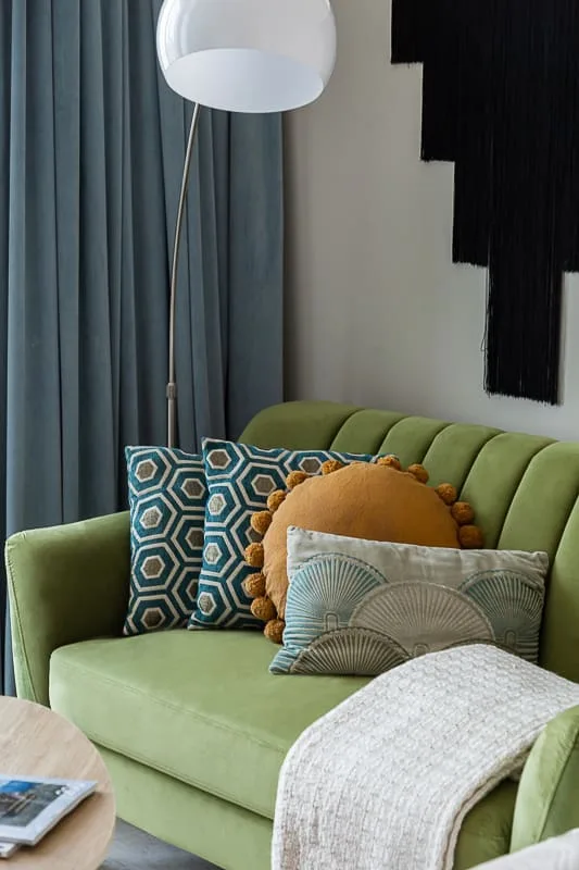 Un sofá verde en una sala de estar junto a cortinas azules, con cojines estampados y una manta.