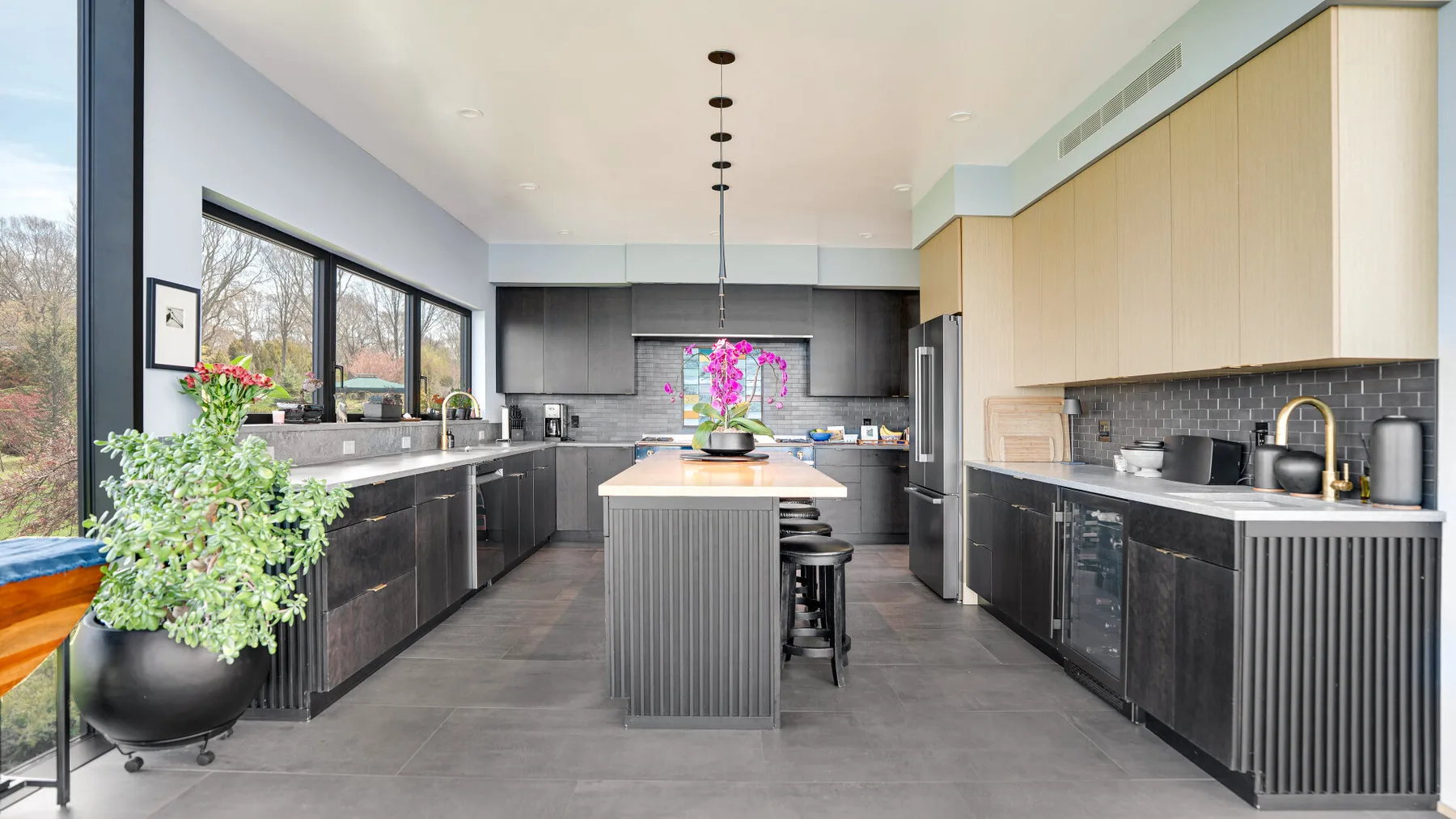 A modern kitchen has dark cabinetry, a central island, and large windows.