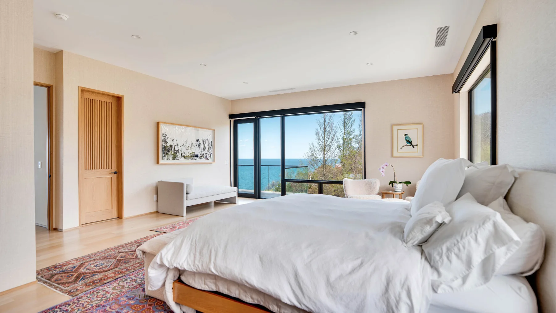 A bright bedroom with a large bed facing wide windows overlooking the water.