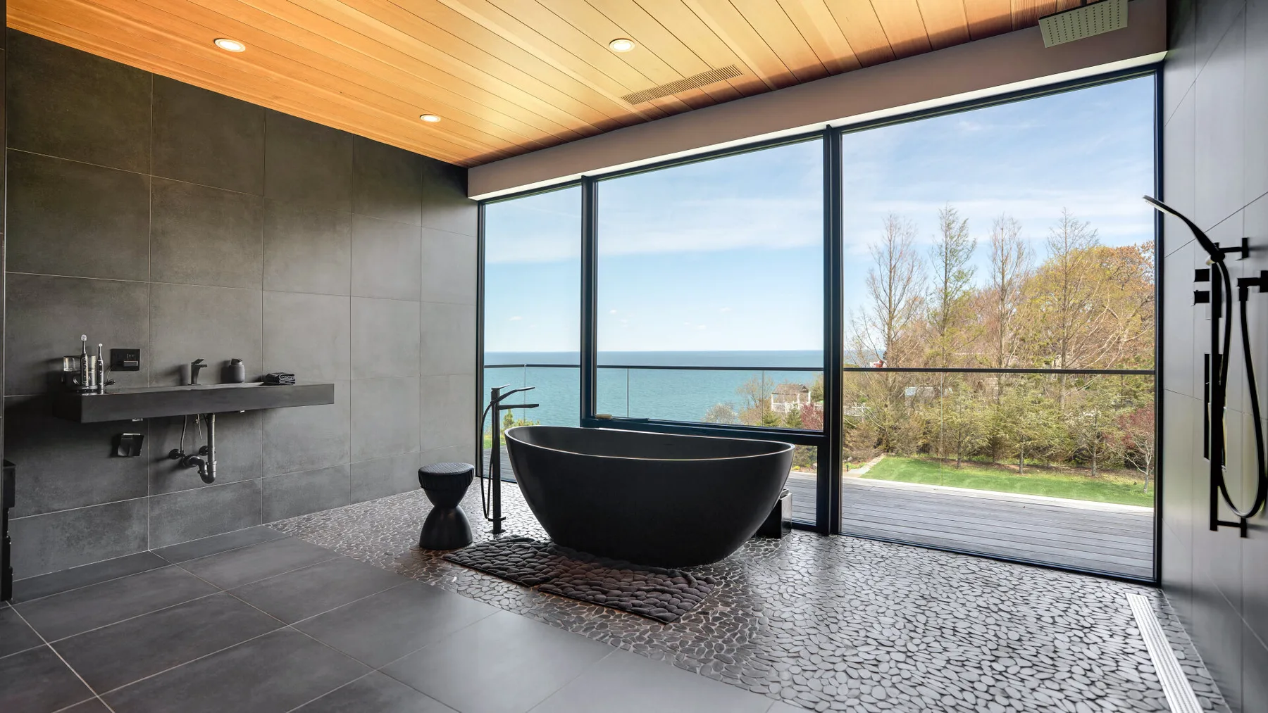 A modern bathroom features a freestanding tub beside large windows overlooking trees and water.