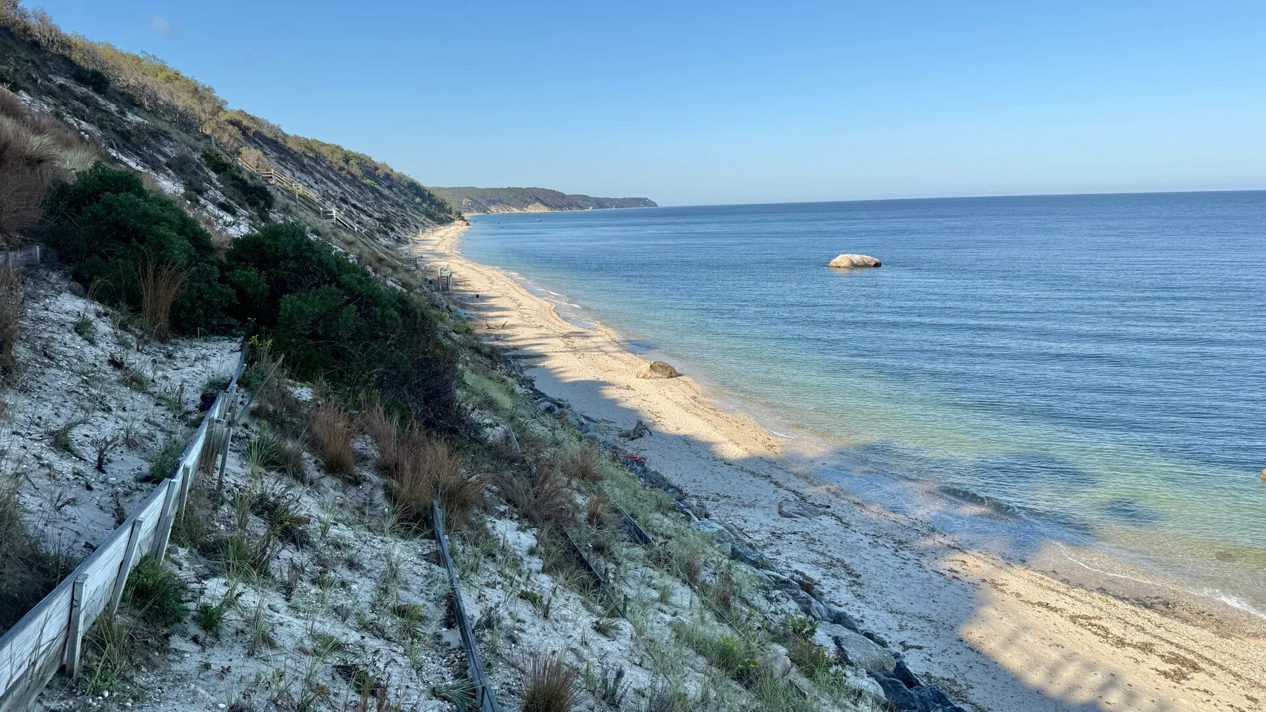 A sandy beach curves along a vegetated coastal bluff beside calm blue water.