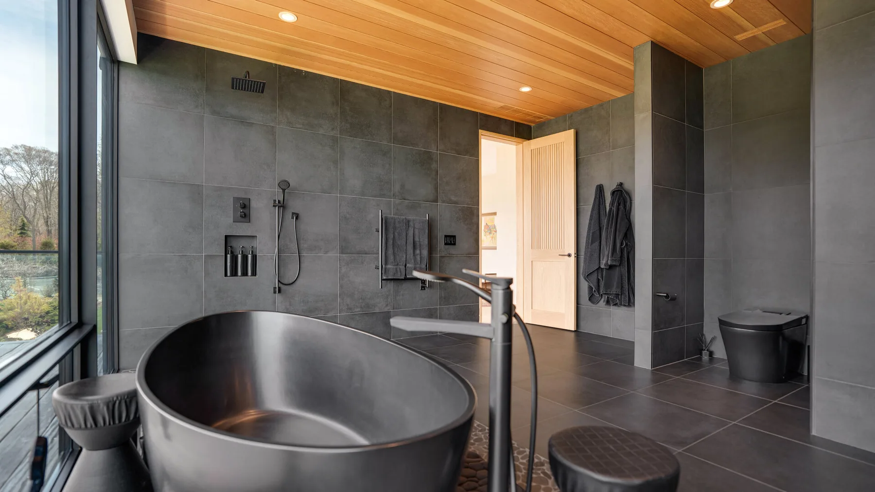 A modern bathroom features a freestanding bathtub, dark tiled walls, and a wood ceiling.