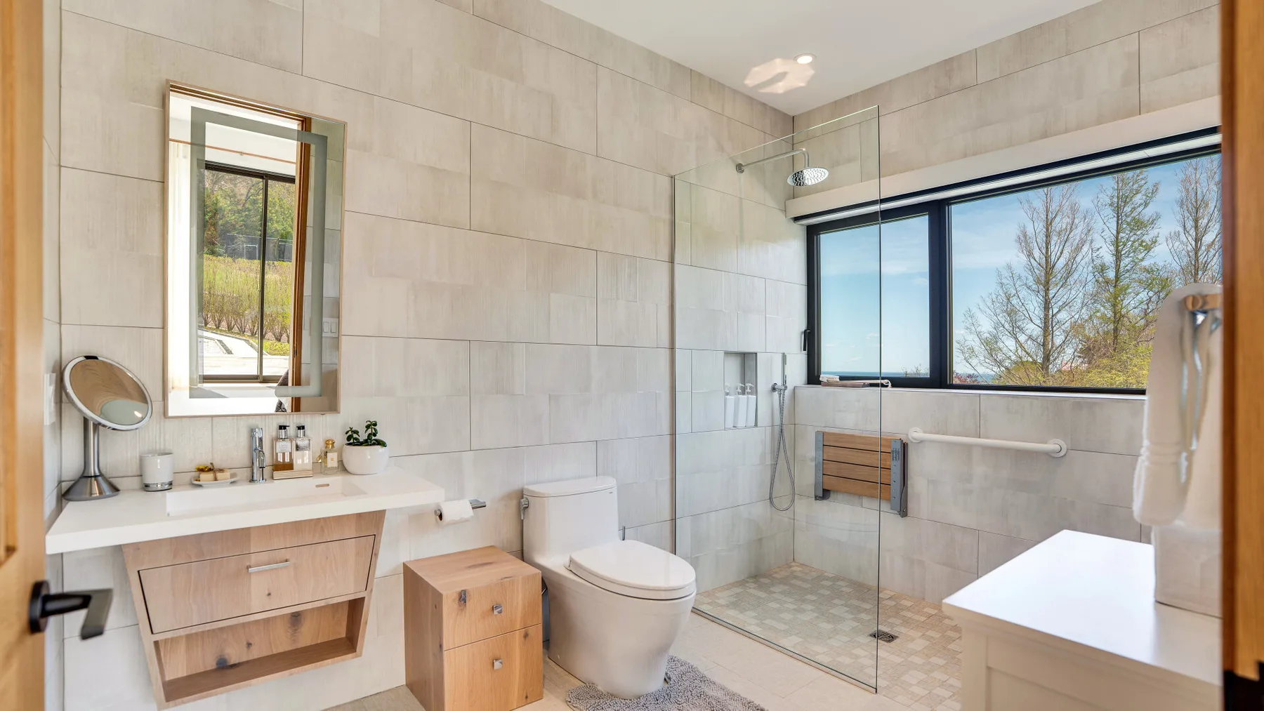 A tiled bathroom with a vanity, toilet, and glass shower beside a large window.