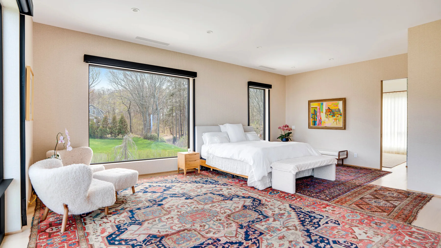 A bright bedroom with a bed, seating, and large windows overlooking trees.