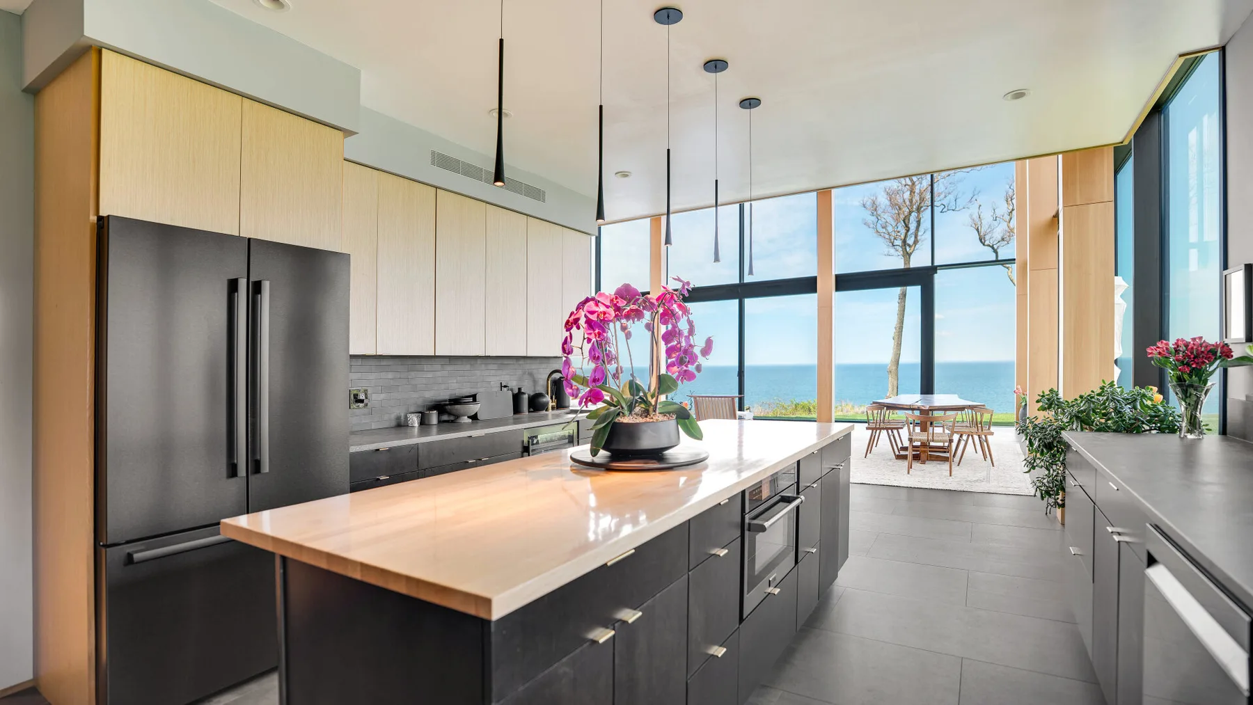 A modern kitchen with a large island opens onto a dining area by floor-to-ceiling windows.