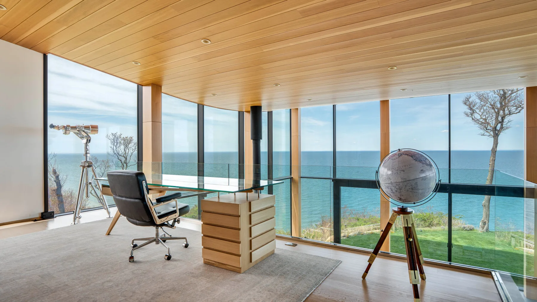A bright room with a desk and telescope beside floor-to-ceiling windows overlooking the sea.