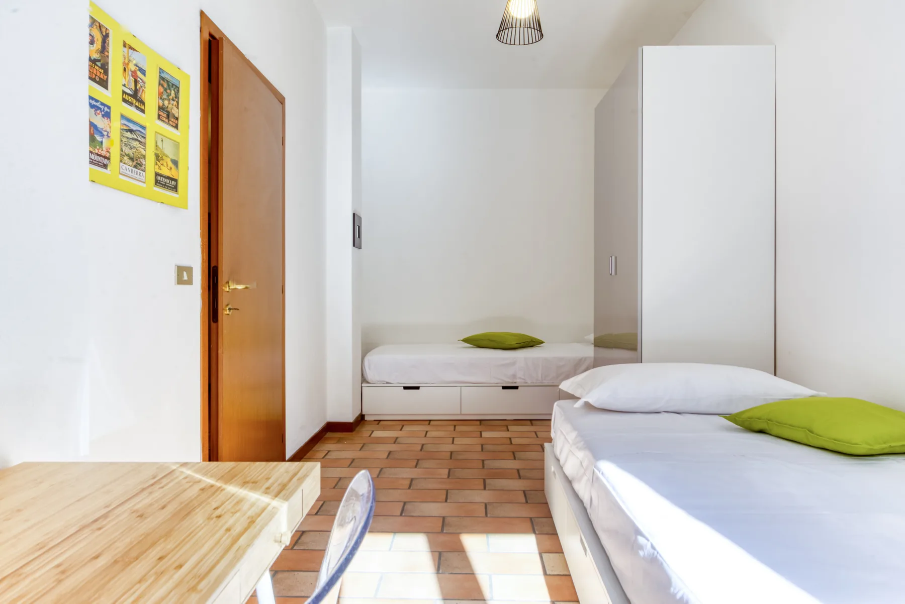 Dormitorio luminoso con dos camas individuales, un armario y suelo de baldosas color terracota.