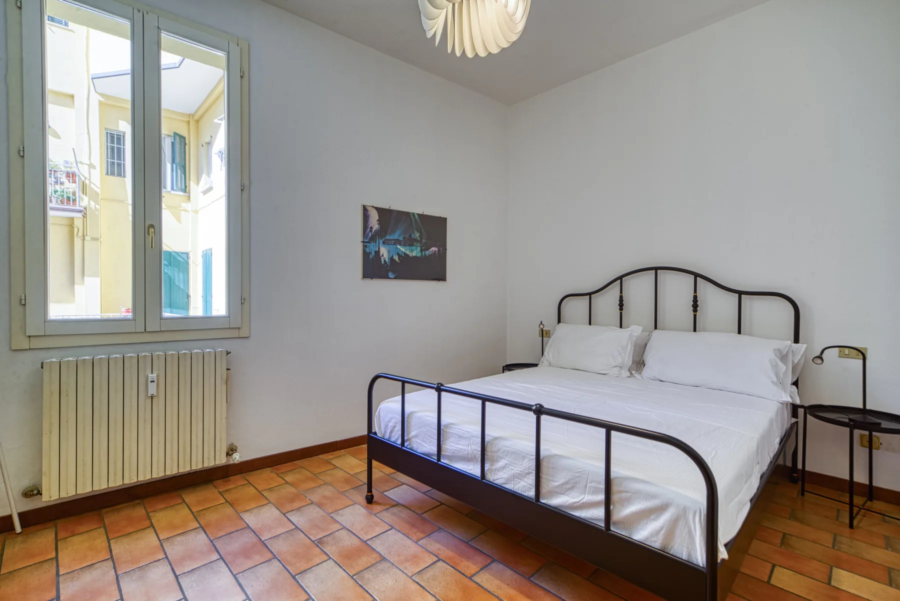 A bedroom with a metal bed, white bedding, and a window overlooking nearby buildings.