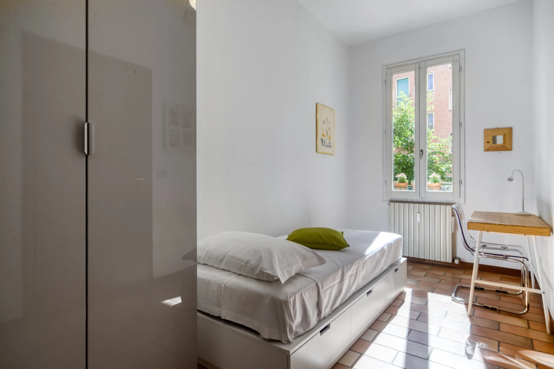 Un dormitorio luminoso con una cama junto a una ventana alta y un escritorio pequeño.
