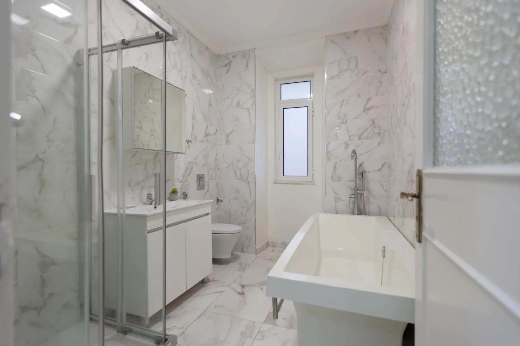 A bright bathroom with a bathtub, glass shower, and white marble-patterned surfaces.