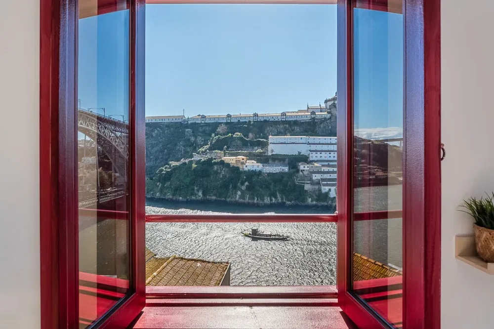A red-framed window overlooks a rocky shoreline and buildings on a hillside.