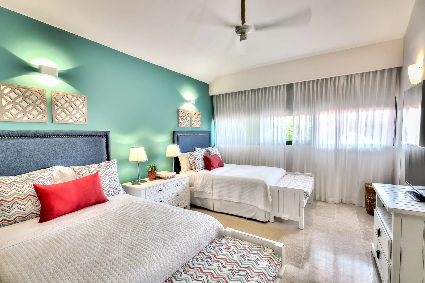 Un dormitorio luminoso con dos camas, cojines de colores y cortinas translúcidas.