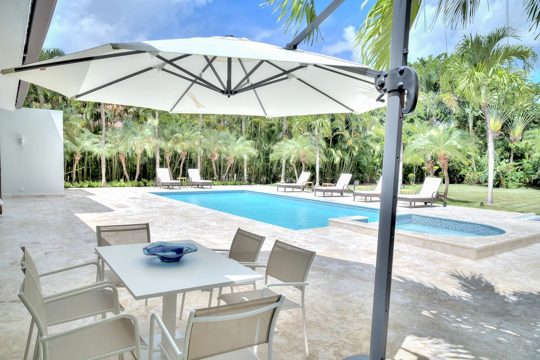 Una piscina al aire libre junto a una mesa y sillas de patio, rodeada de vegetación tropical.