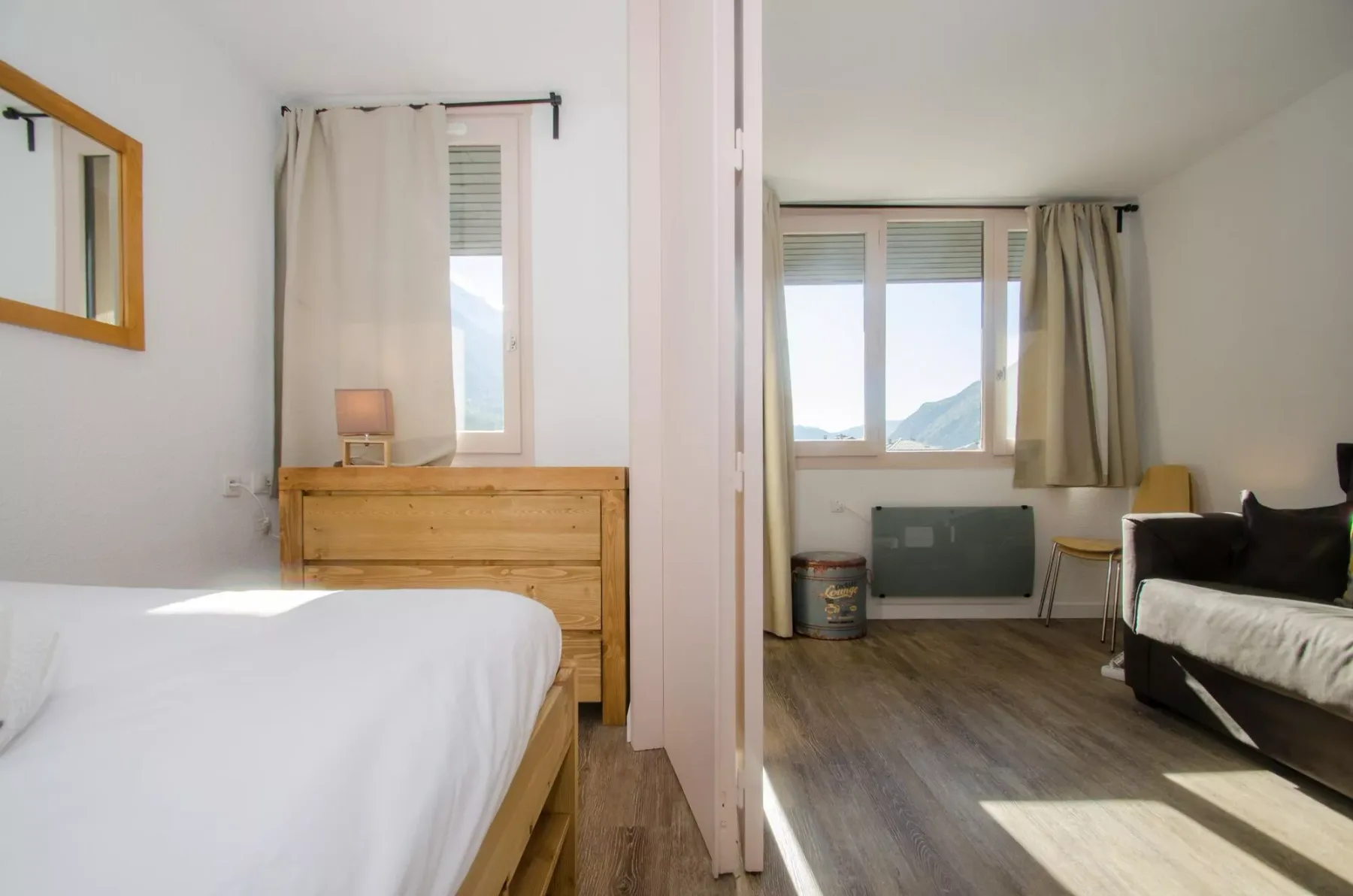 A bedroom with a bed and wooden dresser, opening onto a sitting area by a window.