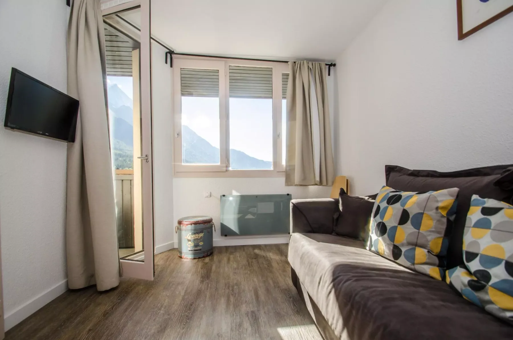 A living room with a sofa, a wall-mounted television, and windows overlooking mountains.