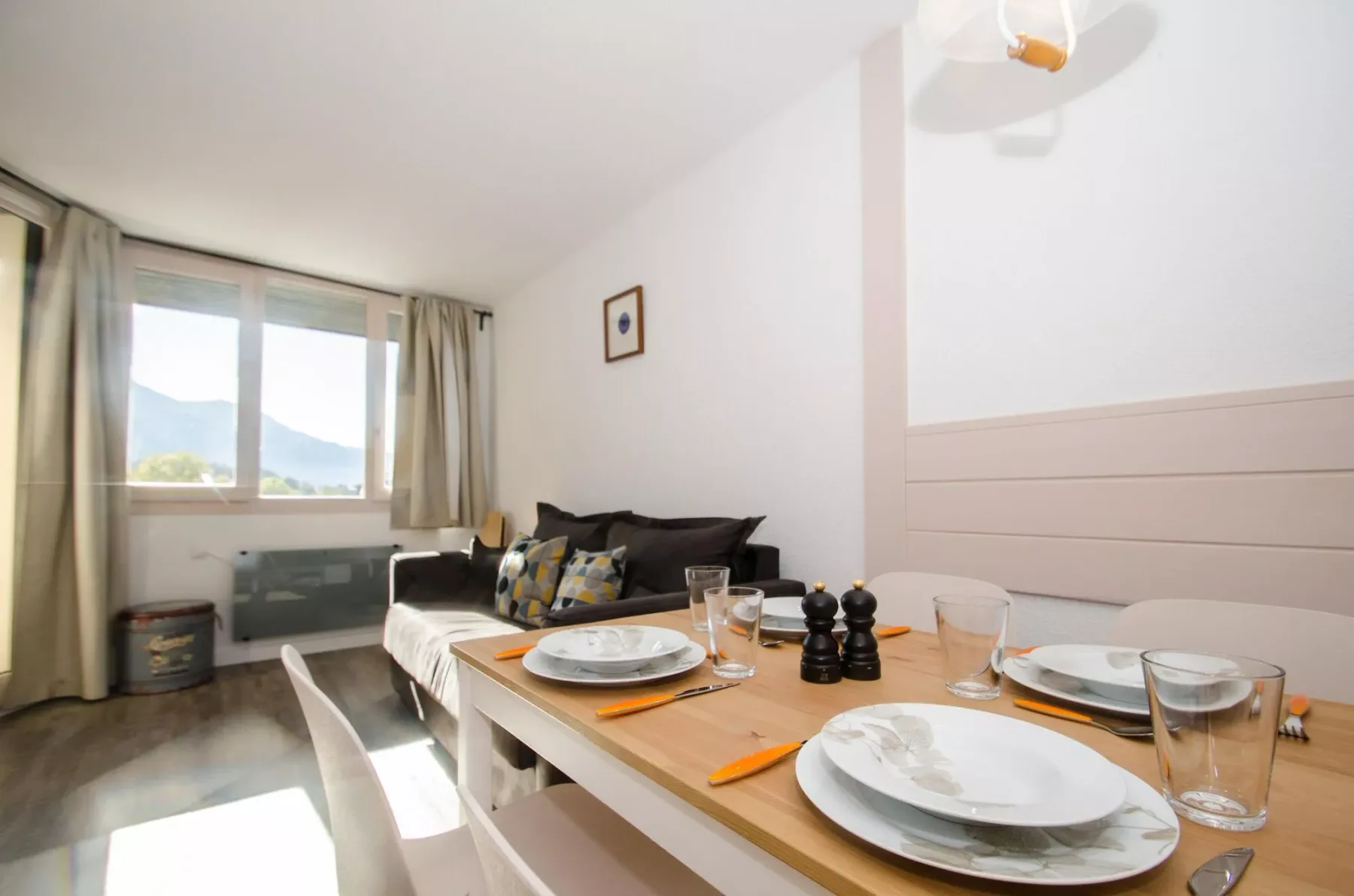 A dining table set with plates and glasses sits beside a living area with a sofa and windows.