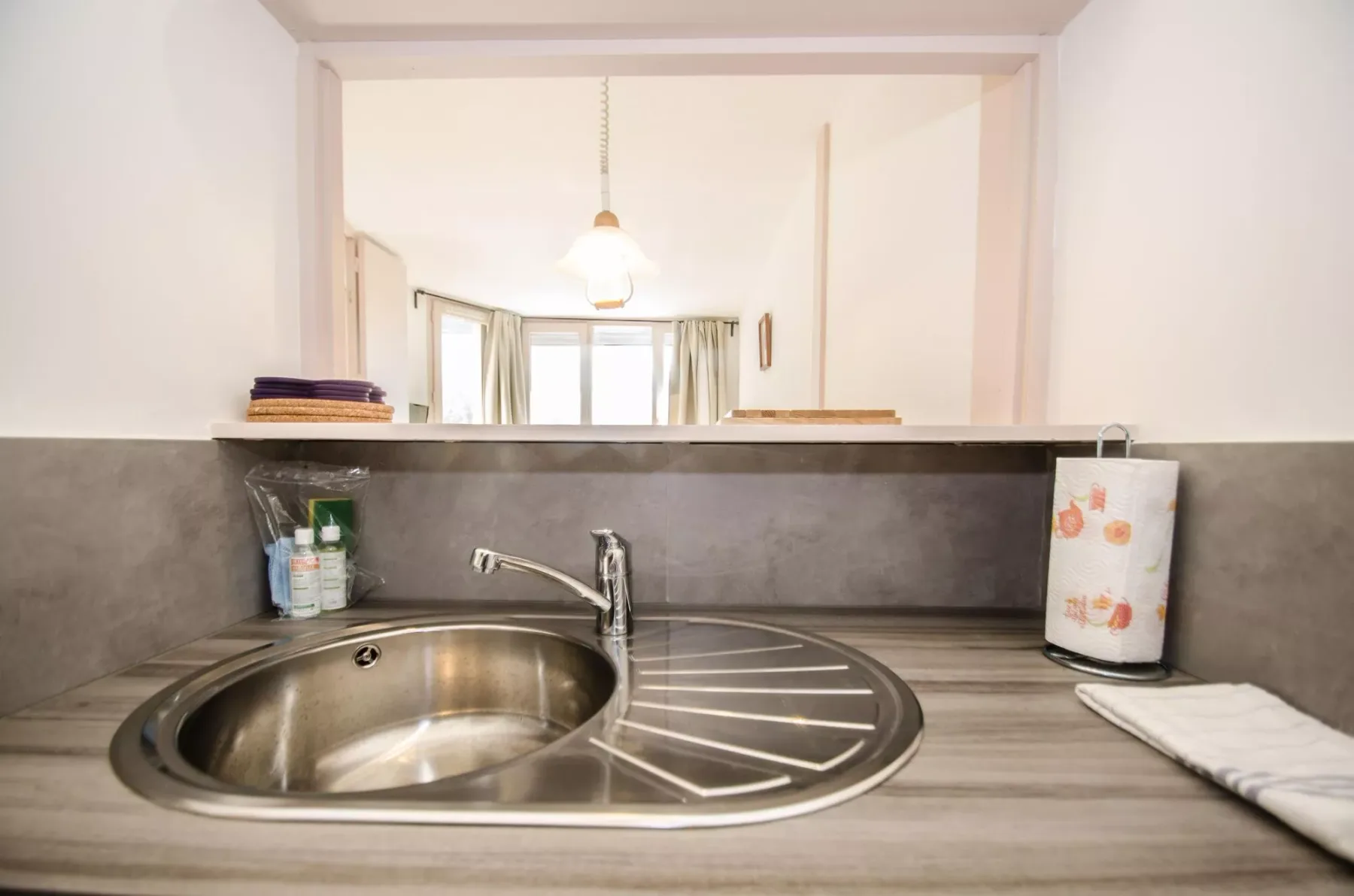 A kitchen sink and drying area sit beneath a wide opening into a bright room.