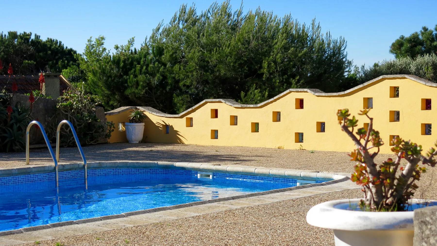 An outdoor swimming pool sits beside a yellow stucco wall and leafy trees.
