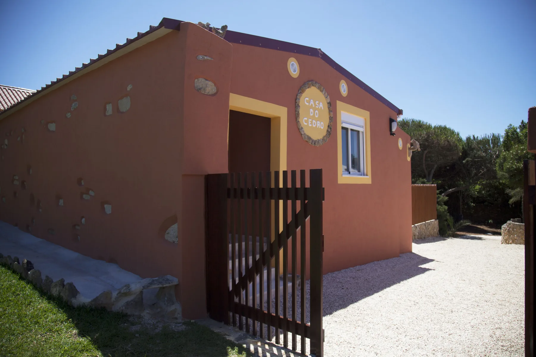 A small reddish building has a dark wooden gate and a window facing a gravel path.