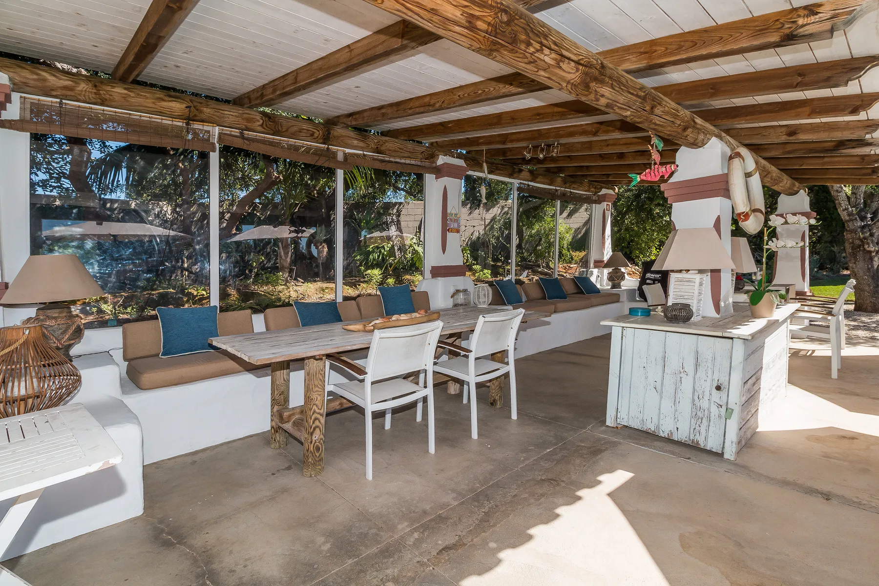 Una zona cubierta para comer al aire libre tiene una mesa larga, asientos y una chimenea integrada.