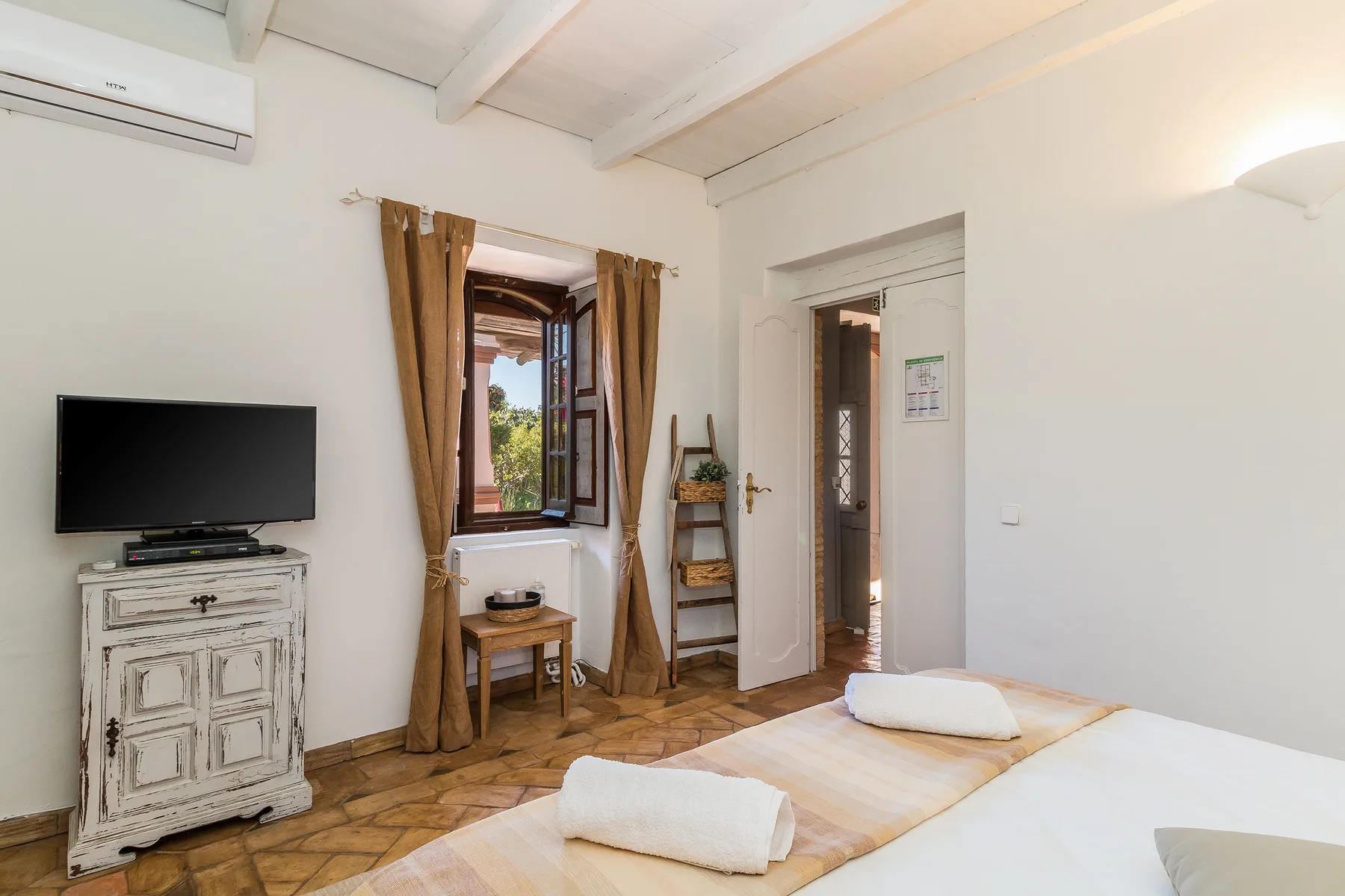 Un dormitorio luminoso con una cama, un televisor y contraventanas de madera abiertas.
