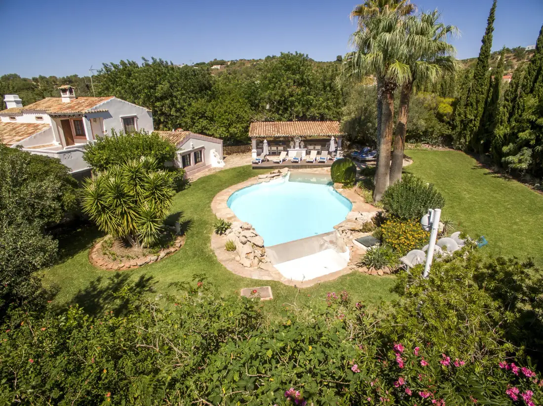 Un jardín rodea una piscina junto a una villa y una zona de estar con sombra.