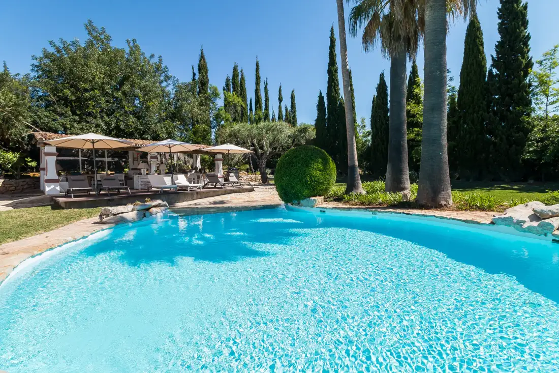 A curved swimming pool sits beside a garden patio with shaded seating.