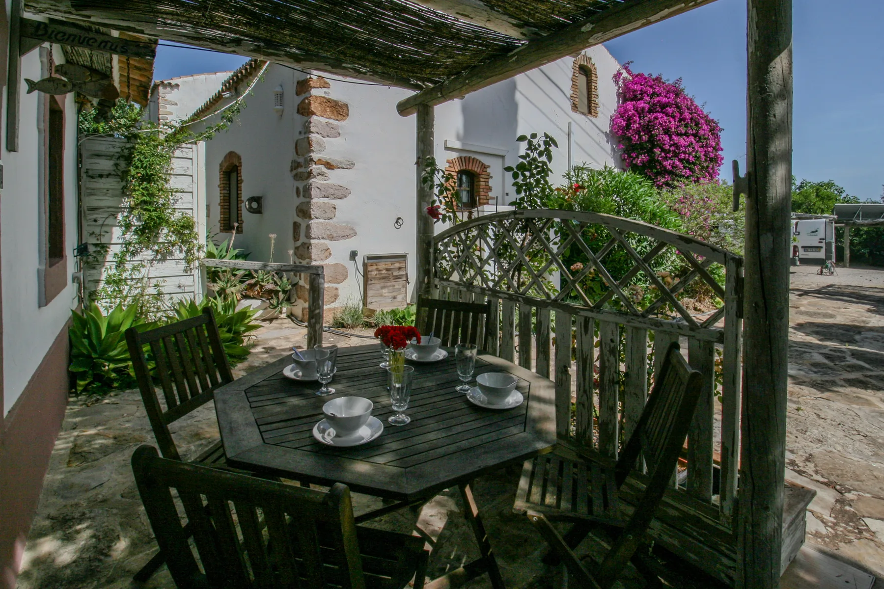 Una zona de comedor exterior a la sombra tiene una mesa y asientos junto a edificios blancos.