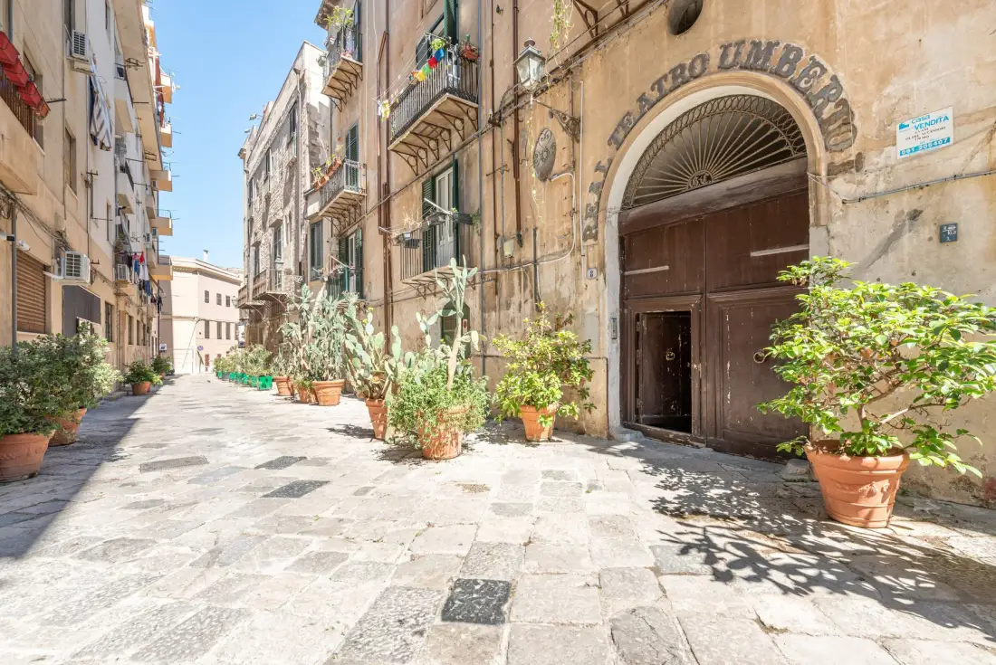 A narrow paved street runs between old buildings, with potted plants along the walls.
