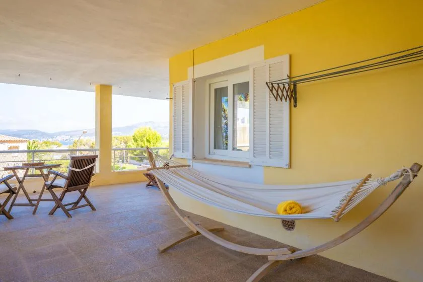 A covered terrace with a hammock, table, and chairs beside a yellow wall.