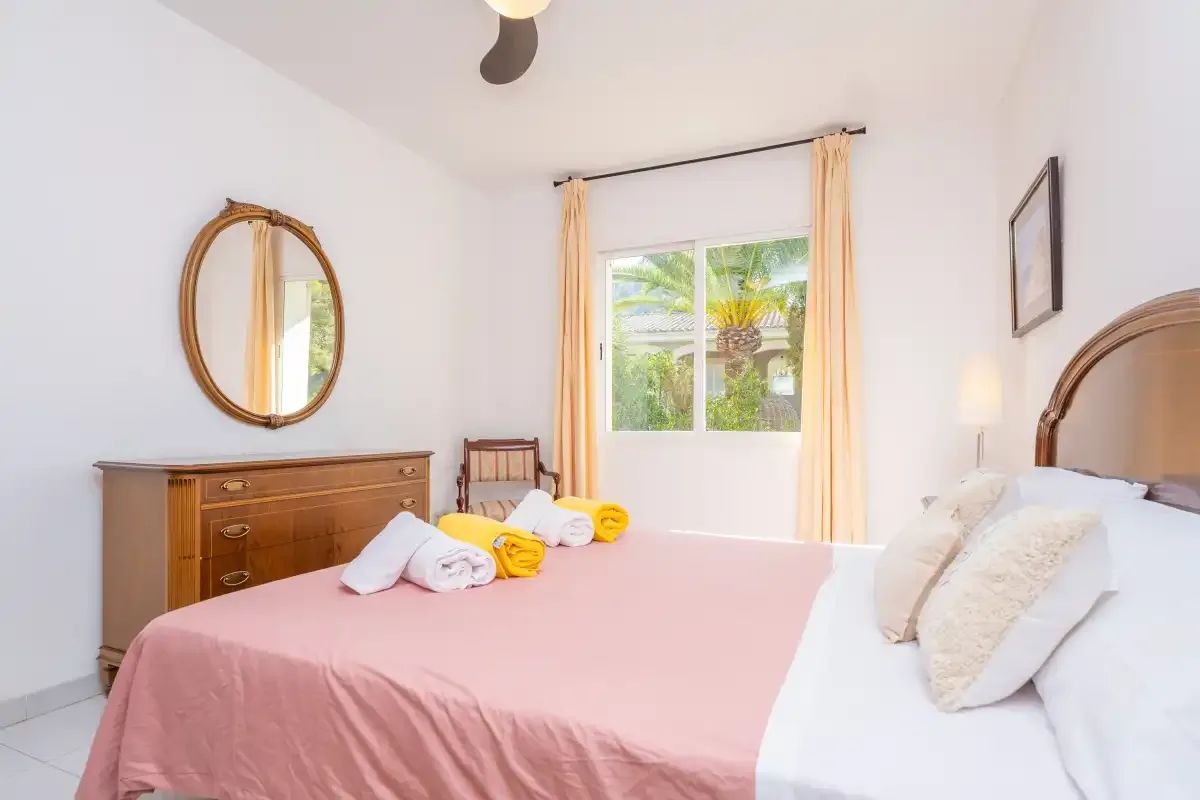 A bedroom with a pink bedspread, wooden dresser, and window overlooking greenery.