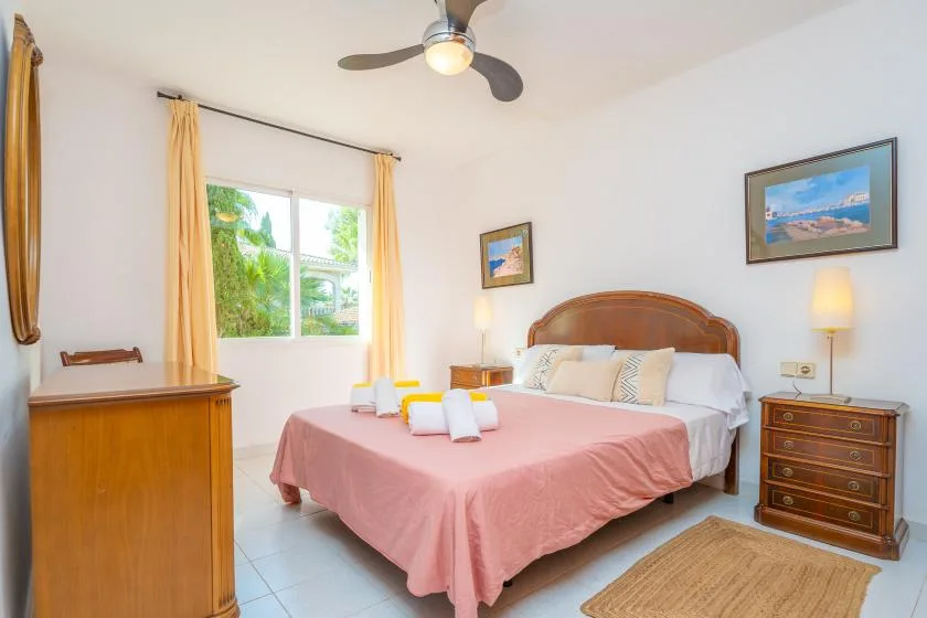 A bright bedroom with a double bed, pink bedspread, and a window with yellow curtains.