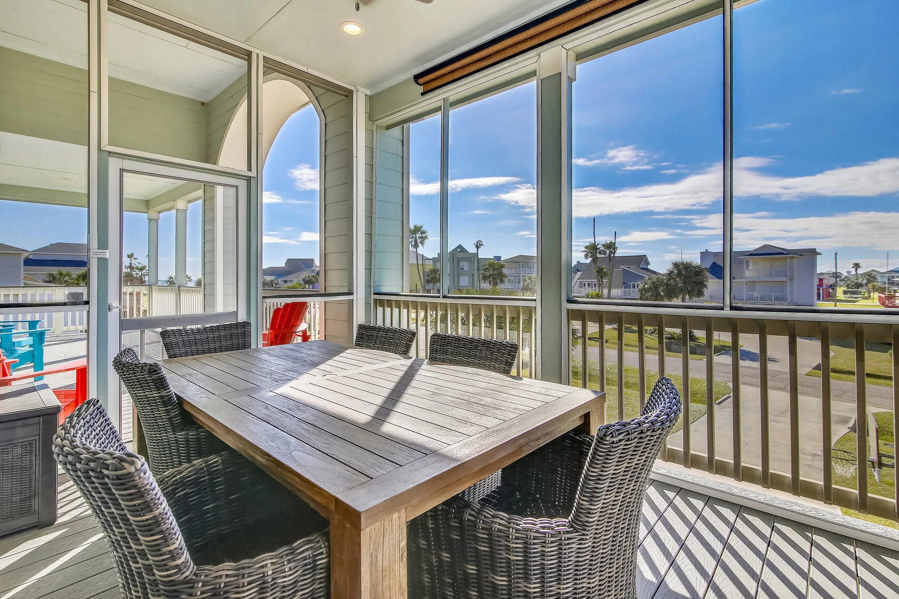 Un comedor exterior cubierto tiene una mesa de madera y asientos tejidos con vistas al vecindario.