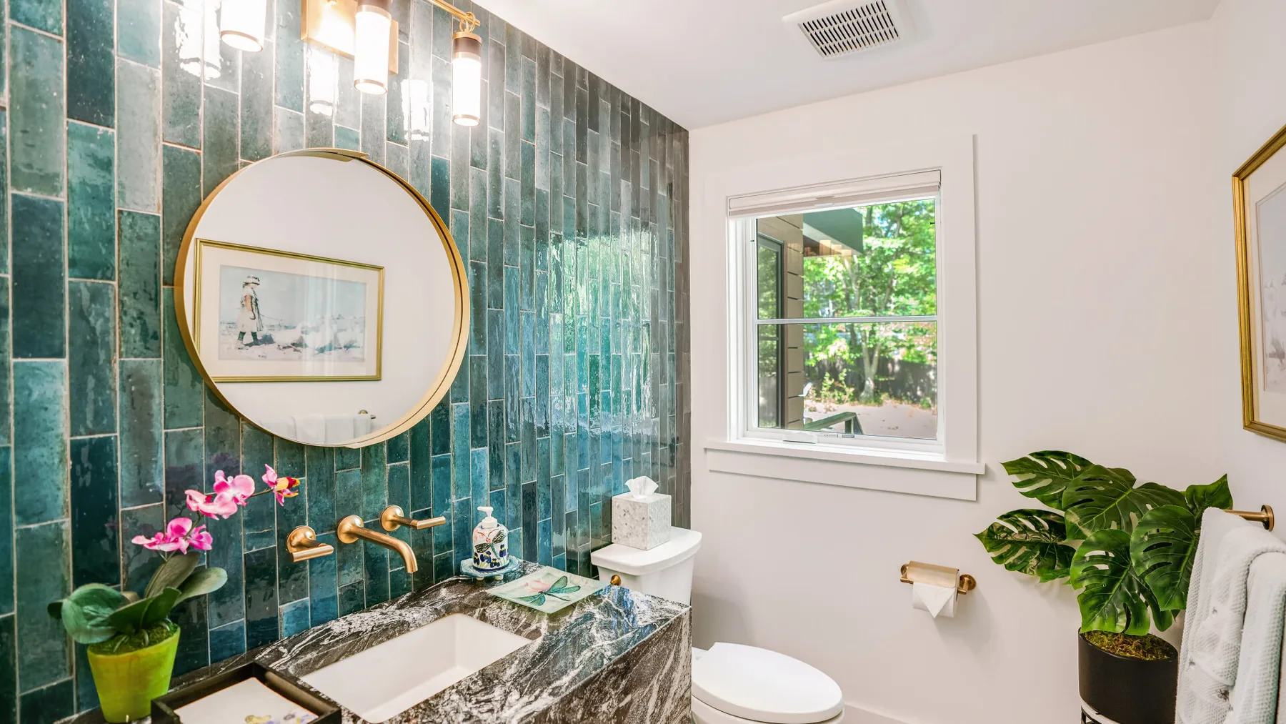 A bathroom with a tiled vanity, round mirror, window, and toilet.