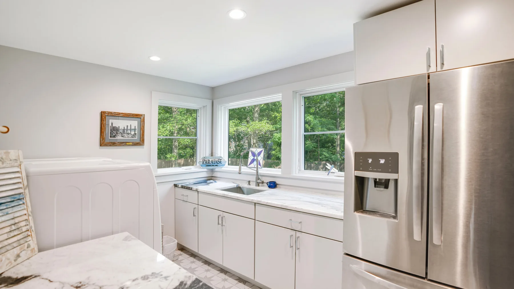 A bright kitchen with a sink beneath large windows and a stainless steel refrigerator.