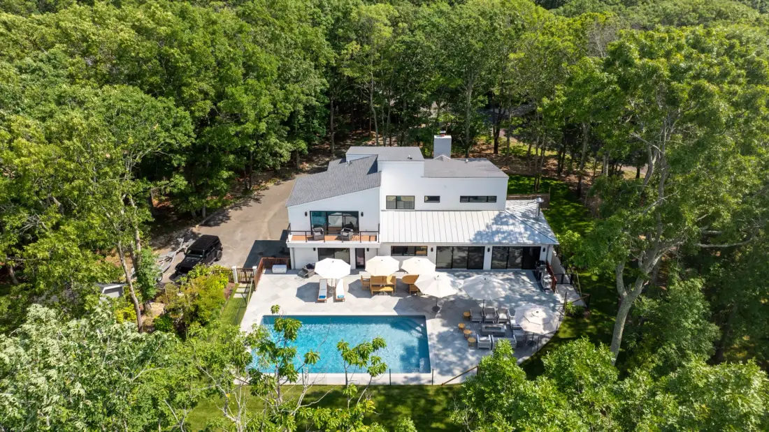 A modern white house sits among trees beside a swimming pool and patio.